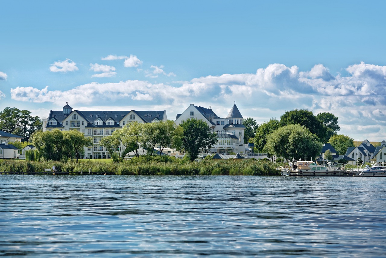 Elegantes Hotel am Wasser, von einem See aus unter blauem Himmel mit flauschigen Wolken betrachtet, umgeben von üppigem Grün.