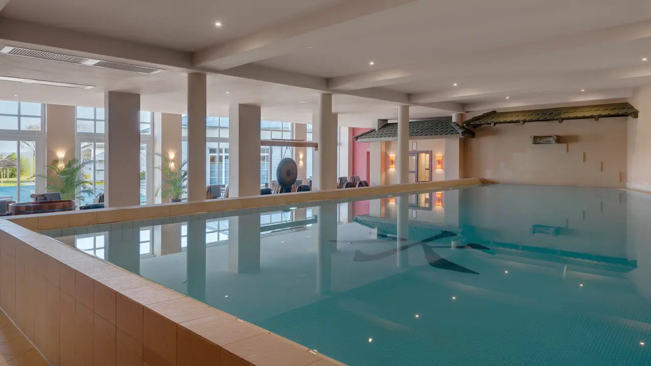 Indoor pool area of a spa with reflective water, surrounded by columns and relaxation lounges, offering a tranquil environment.