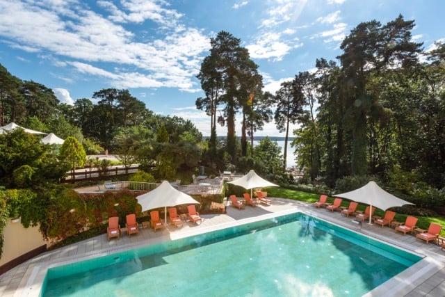 Outdoor pool surrounded by umbrellas and sun loungers with a dense forest backdrop at the Precise Resort Bad Saarow.