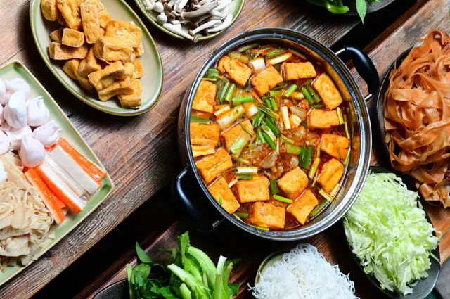 Asiatisches Hotpot-Gericht mit Tofu und Gemüse auf einem Holztisch umgeben von verschiedenen Beilagen.