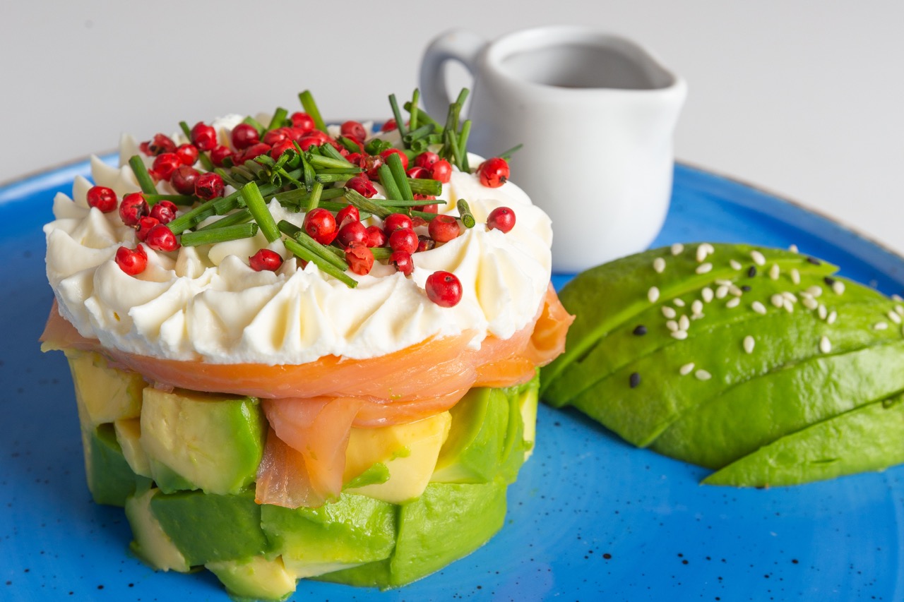 An artistically arranged avocado salmon tartare, topped with cream and fresh herbs, served on a blue plate.