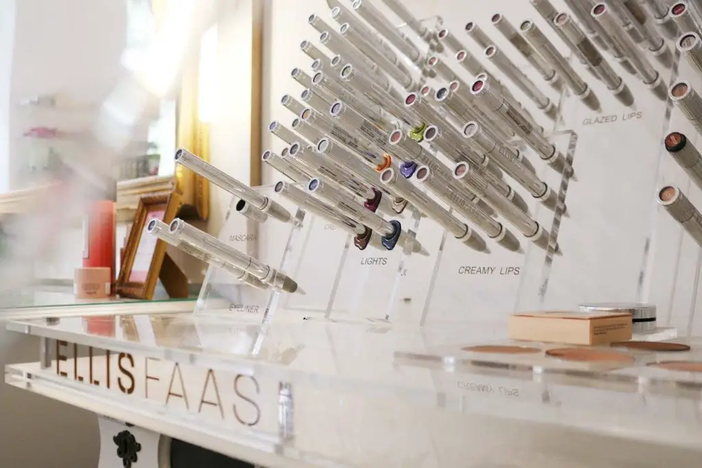 Eine Auswahl von ELLIS FAAS Make-up-Produkten auf einem klaren Acrylständer mit Markenlogo, vor einem verschwommenen Salon-Hintergrund.