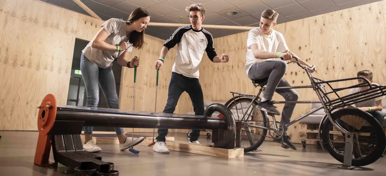 Personen spielen eine kreative Variante von Minigolf mit einem Fahrrad auf einer Holzbahn.