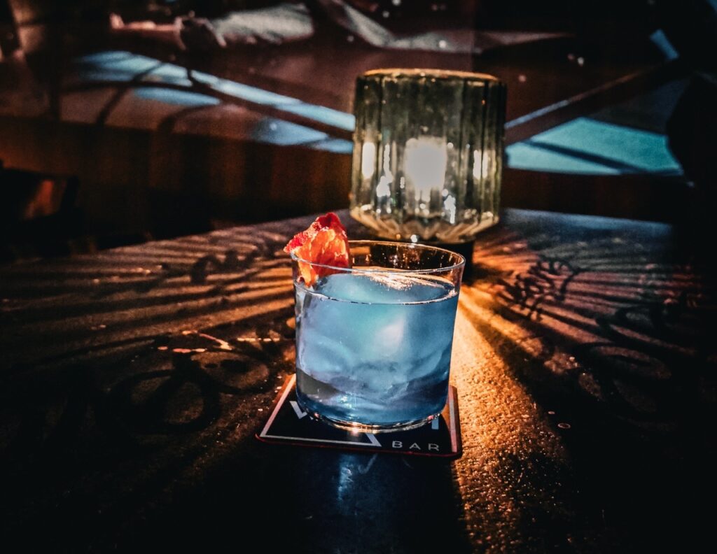 This photograph captures a quintessential scene at the Wash Bar in Berlin Mitte: a finely crafted cocktail awaiting the first sip. The subtle glow of candlelight bathes the drink in a warm, inviting light, casting intricate shadows across the table that speak to the bar’s chic and intimate ambiance. With a garnish of vibrant orange …