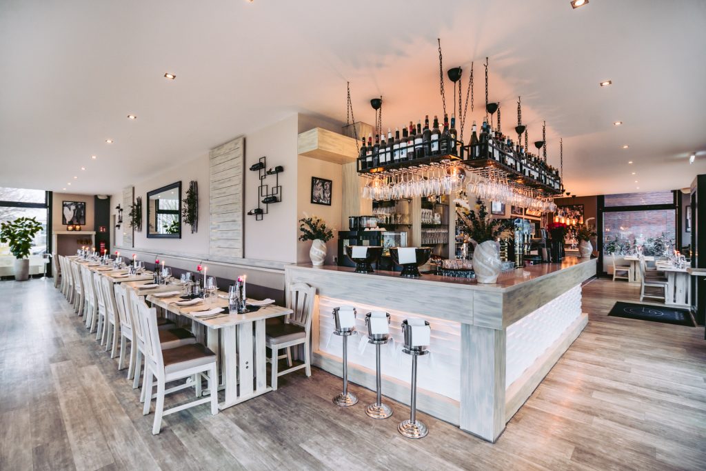 Blick in den gesamten Restaurantbereich mit langer eingedeckter Tafel, Bar mit Hängeregal für Gläser und Flaschen, modern-hell eingerichtet im griechischen Restaurant Greek Food Olympia.