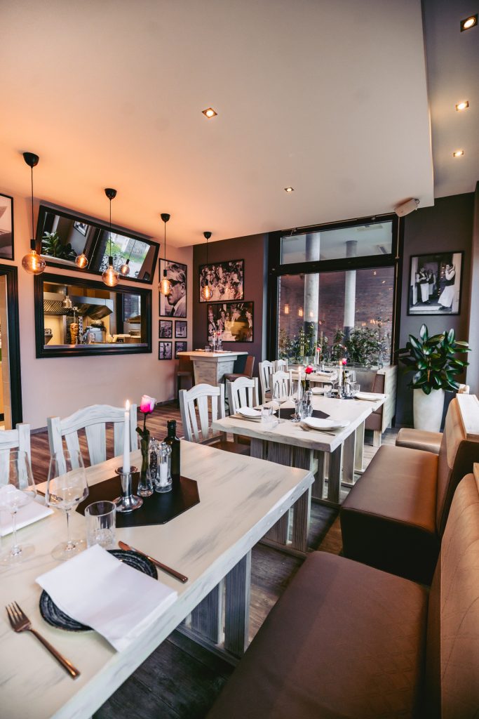 Gemütlicher Gastraum mit hellen Holztischen, romantisch gedeckt mit Kerzen und Gläsern, im Hintergrund große Fenster und Fotos an der Wand im griechischen Restaurant Greek Food Olympia.