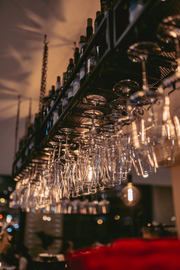 Nahaufnahme einer beleuchteten Bar, an der zahlreiche Weingläser kopfüber hängen, mit Flaschen oben auf der Ablage im griechischen Restaurant Greek Food Olympia.