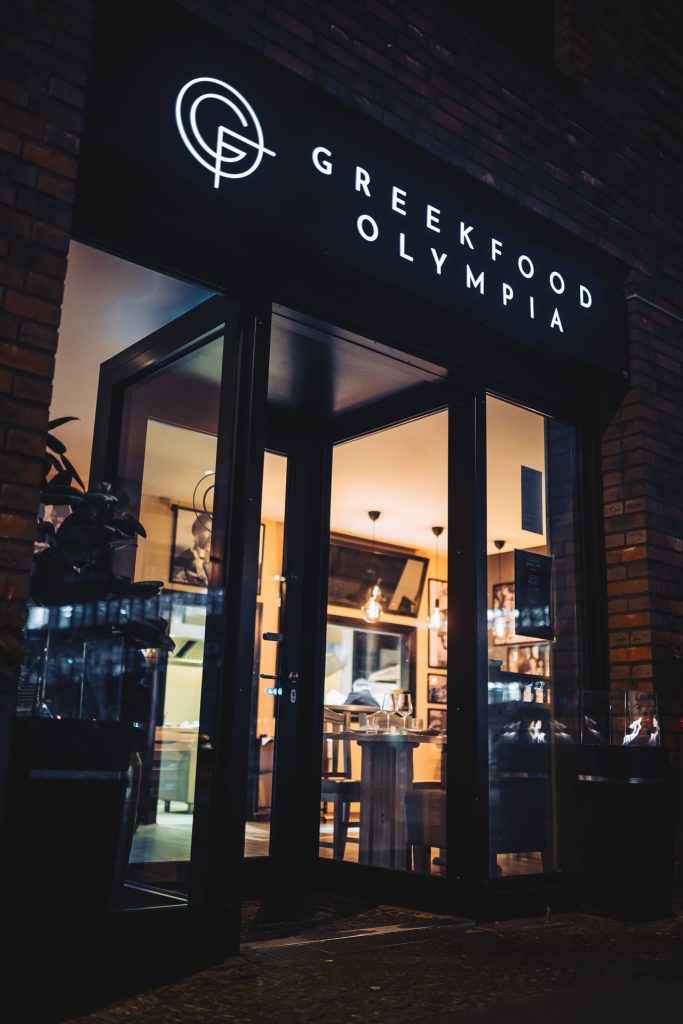 Außenansicht des Eingangs von „Greek Food Olympia“ mit beleuchtetem Schriftzug über der Tür, Blick hinein in das stimmungsvolle Innere im griechischen Restaurant Greek Food Olympia.
