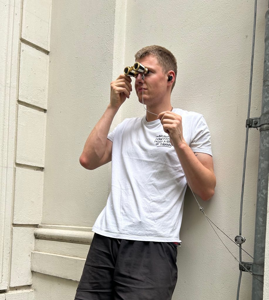 Mann blickt konzentriert durch ein Opernglas an einer Hauswand, mit Kopfhörern verbunden.