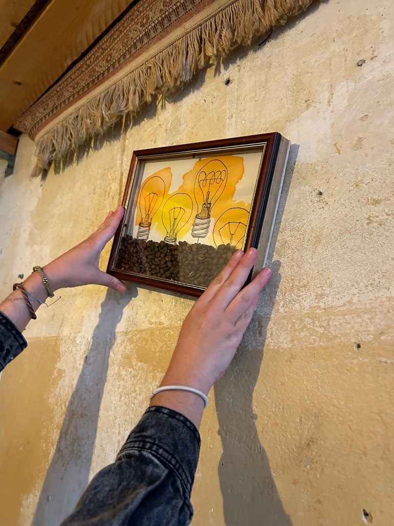 Nahaufnahme: Zwei Hände hängen ein gerahmtes Bild mit Glühbirnen und Kaffeebohnen an eine rustikale Wand.