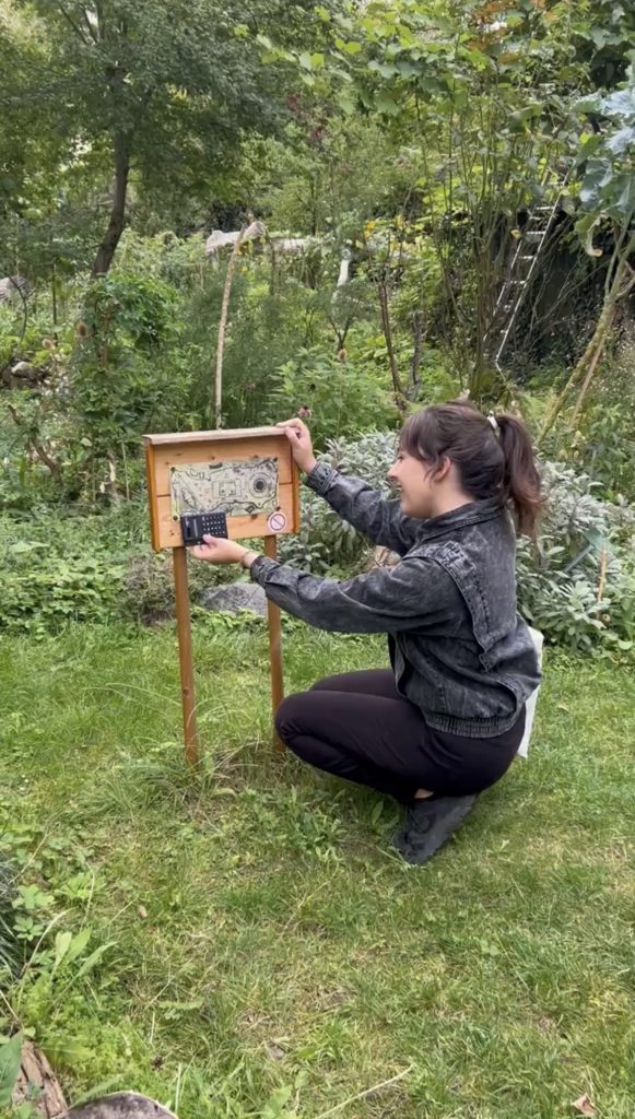 Junge Frau kniet in einem grünen Garten und öffnet ein Zahlenschloss an einer Holzstation.