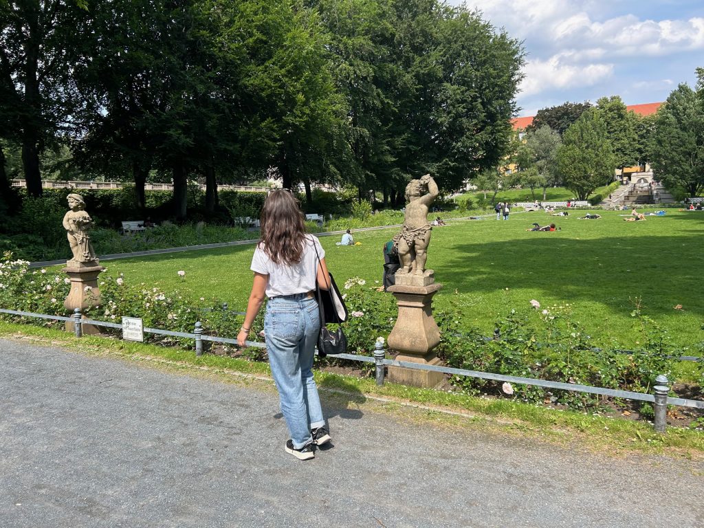 Frau spaziert durch eine gepflegte Grünanlage mit Statuen, während andere Menschen im Hintergrund auf der Wiese entspannen.