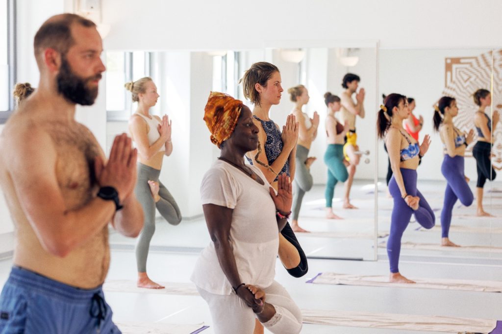 Diverse Gruppe von Menschen übt Baum-Yoga im hellen Mahalaya Yoga & Healing Arts Studio.